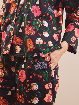 A close-up of a person wearing a matching pyjama set with a black background and a colourful floral print. The print features pink, red, orange, green, and white flowers with green stems and leaves.