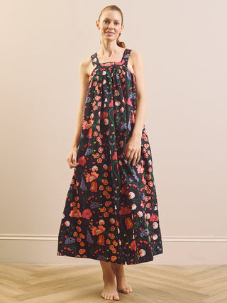 A woman modeling a sleeveless black maxi nightgown with a colorful floral and butterfly print, standing barefoot on a light wood floor against a beige wall.
