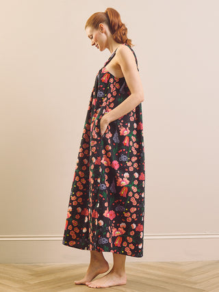 A woman with red hair tied back is shown from the side, wearing a sleeveless black maxi nightdress with a colourful floral print. She is standing barefoot on a light wood floor against a plain beige wall.