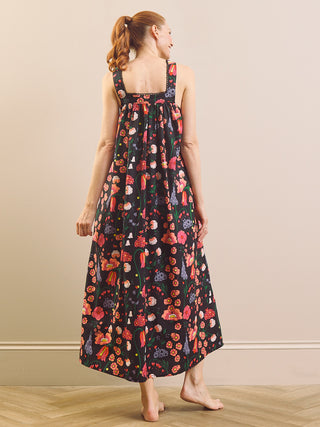 A woman stands barefoot indoors wearing a sleeveless maxi nightdress with a dark background and an allover colourful floral and fruit print. She is photographed from the back against a plain beige wall and light wood floor.