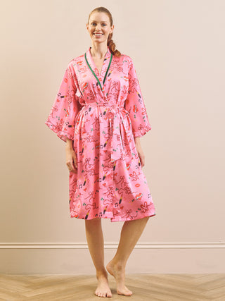 A woman modelling a pink silk-like robe with a playful multicolour print of cartoon characters, lips, and hearts, featuring green trim on the V-neck. She is standing barefoot on a light wood floor against a beige wall.