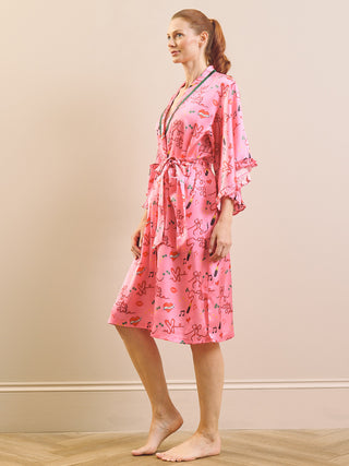 A woman wearing a pink satin-like robe with a playful multicolor cartoon print, standing barefoot against a neutral beige wall.