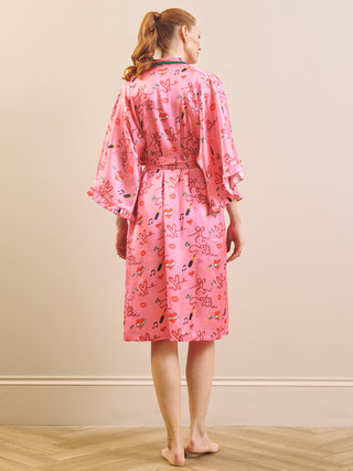 A woman stands barefoot on a light wood floor against a beige wall, viewed from the back, wearing a pink satin-like robe with a multicolor floral and abstract print.