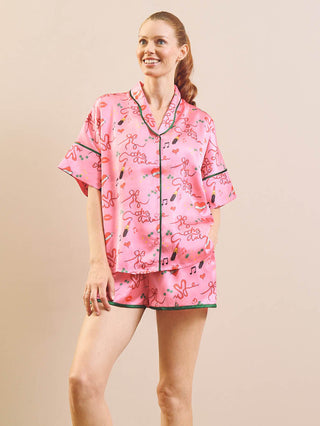 A woman modelling a pink short-sleeve pyjama set with a playful cartoon-style print and dark green piping, set against a solid beige background.