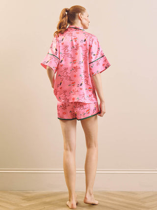 A woman stands barefoot indoors wearing a pink short-sleeve pajama set with a cartoon print and dark green piping, photographed from the back against a plain beige wall.