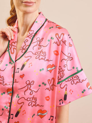 A close-up of a woman wearing a pink satin pyjama top with a graphic print of lipstick kisses, hearts, music notes, and the words 'cheer lifestyle' in brown script, accented with dark green piping.