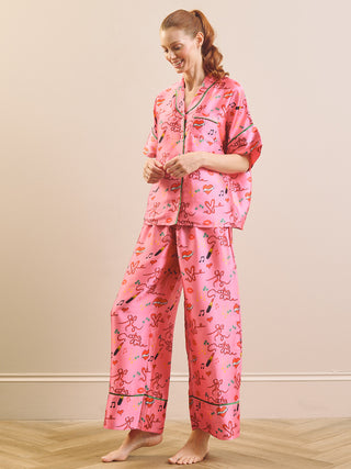 A woman modelling a pink pyjama set with a colourful musical note and whimsical character print, standing against a neutral beige wall.