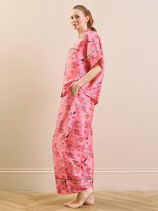 A woman modelling a pink short-sleeve pyjama set with a playful multicolour cartoon print, standing against a beige wall and light wood floor.