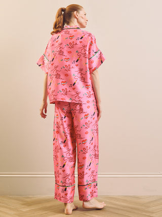 A woman stands barefoot indoors wearing a pink pyjama set with a playful multicolour print of cartoon characters, hearts, and lips, photographed from the back against a plain beige wall and light wood floor.
