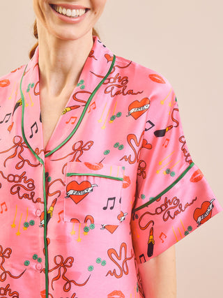 A close-up of a woman wearing a pink short-sleeve pyjama shirt with a colourful graphic print of hearts, lips, musical notes, and script text, featuring green piping.