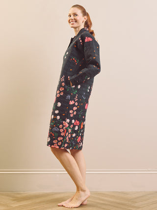 A woman modelling a long quilted house coat dress with a dark navy base and a colourful floral patchwork print, standing barefoot on a light wood floor against a beige wall.