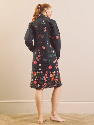 A woman stands barefoot indoors, viewed from the back, wearing a dark quilted house coat robe with a colourful floral print. The robe has a textured, stitched quilted pattern and features pink, red, orange, and white flowers with green stems.