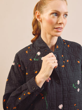 A woman wearing a black quilted house coat with embroidered orange, pink, and green floral details, shown in a close-up shot against a solid beige background.