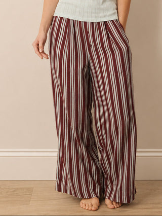 A woman stands barefoot against a beige wall wearing a light sage green ribbed tank top and wide-leg pants with vertical burgundy and cream stripes.