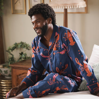 Man wearing blue pajamas with red lobster pattern sitting on a bed in a bedroom.