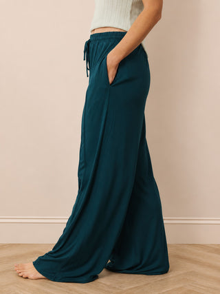 A person wearing loose, wide-leg teal pants with an elastic drawstring waist and side pockets, standing barefoot on a light wood floor against a beige wall.