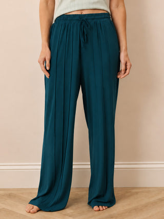 A person wearing loose, wide-leg teal drawstring trousers standing barefoot on a light wood floor against a beige wall.