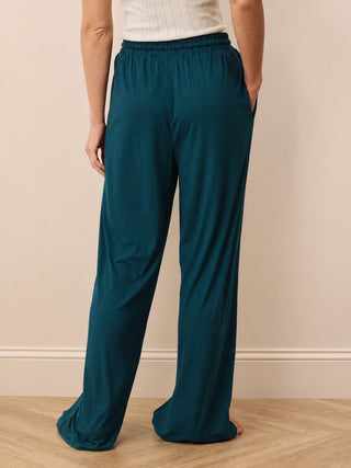 A person shown from the back wearing loose, wide-leg teal pants with an elastic waistband, standing against a beige wall on a light wood floor.