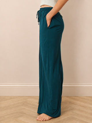 A person wearing loose teal drawstring lounge pants, shown from the waist down in a side profile against a beige wall and light wood floor.