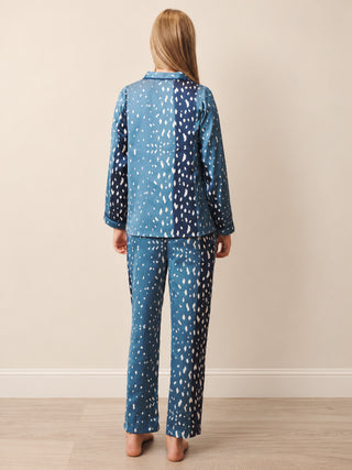 A girl stands with her back to the camera wearing a matching two-piece pyjama set with a blue and white abstract spotted pattern, set against a neutral beige wall and light wood floor.