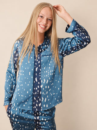 A smiling blonde girl wearing a matching two-piece pyjama set in blue with a white speckled pattern, posed against a solid beige studio background.