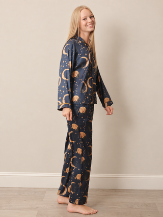 A woman modelling a two-piece pyjama set with a dark navy blue background and a celestial pattern of gold crescent moons, stars, and planets, standing against a beige wall and light wood floor.