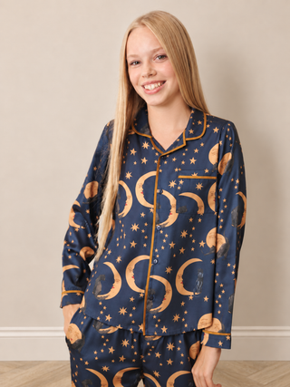 A smiling young woman wearing a navy blue pyjama set with a gold celestial print of crescent moons, cats and stars, standing against a neutral beige wall.
