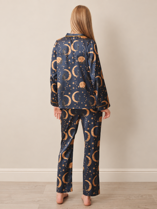 A woman viewed from the back wearing a matching pyjama set with a navy blue satin-like fabric and an all-over gold celestial print of crescent moons, stars, and planets, standing against a plain beige wall.