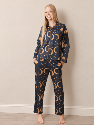 A woman modelling a two-piece pyjama set with a dark navy blue background and a gold celestial print of crescent moons and stars, standing against a neutral beige wall.