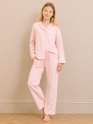A young girl modeling a pink and white vertically striped pajama set, consisting of a long-sleeve button-up shirt and matching pants, standing against a neutral beige wall.
