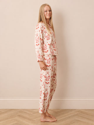 A woman modelling a two-piece pyjama set with a pink bow and ribbon pattern on a cream background, standing against a beige wall and light wood herringbone floor.