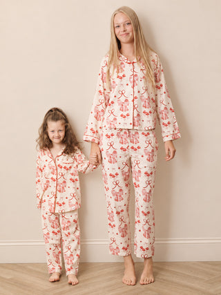 A teenager and a young girl wearing matching pyjama sets with a pink and red gift box pattern on a cream background, standing barefoot against a beige wall.