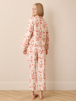 A teenager stands with her back to the camera wearing a matching two-piece pyjama set with a pink and red bow pattern on a cream background, set against a neutral beige wall and light wood floor.