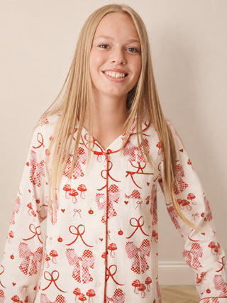 A smiling young girl with long blonde hair wearing a cream-colored long-sleeve pyjama top with a red mushroom and bow pattern, standing against a plain beige wall.