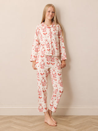 A teenage girl stands barefoot against a beige wall wearing a matching pyjama set with a white base and a red bow and ribbon pattern.