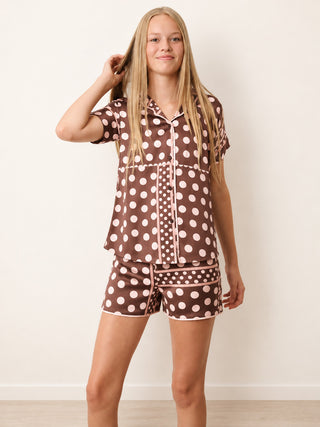 A woman modeling a matching short-sleeve button-up pyjama shirt and shorts set in brown with a white polka dot pattern, set against a plain off-white wall.