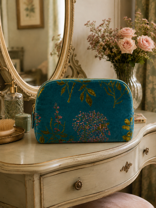 Blue floral-patterned makeup bag on a vintage vanity table with a mirror and flowers in the background.