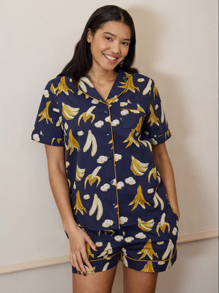 A woman standing, wearing navy blue cotton shortie pyjamas with a hand-drawn banana pattern.