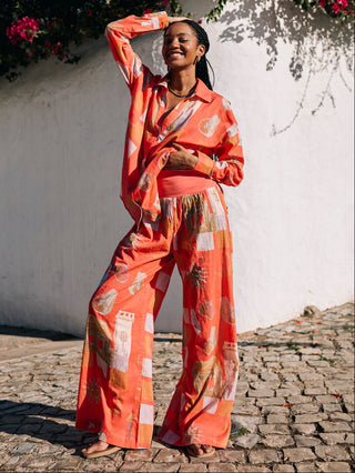 A woman standing on a cobblestone street against a white wall with terracotta-colored floral print trousers and a matching top.