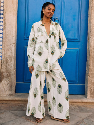 A woman standing in front of a blue door, wearing white linen mix trousers with a green peacock feather print, paired with a matching shirt.