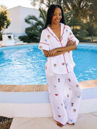 A woman standing by a pool wearing white gauze pyjamas with multicolor embroidery and scallop collar details.