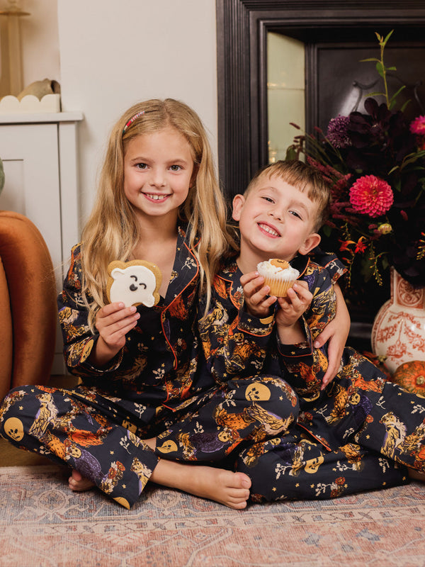 Two children in matching autumn pajamas sitting on the floor in front of a fireplace, holding cookies.