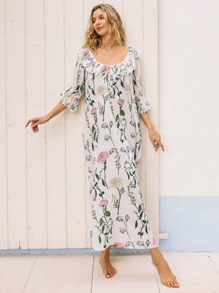 A woman standing in a breezy room wearing a long, floral nightdress with a frilly neckline and sleeves.