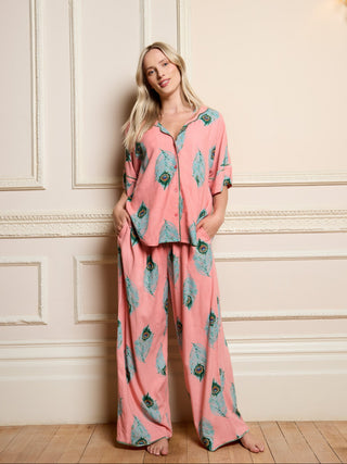Woman wearing a pink peacock pyjama set with peacock print in a room with white paneled walls.