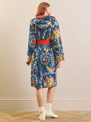 A woman stands with her back to the camera wearing a hooded robe with a blue base and a colorful floral and bird pattern, featuring a red belt and red hood lining, paired with white socks in an indoor setting.