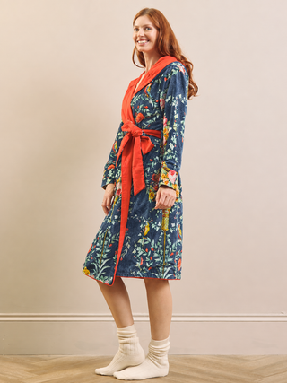 A woman wearing a long-sleeved floral print bathrobe with a bright orange-red collar, tie belt, and lining, paired with cream knit socks, standing against a beige wall.