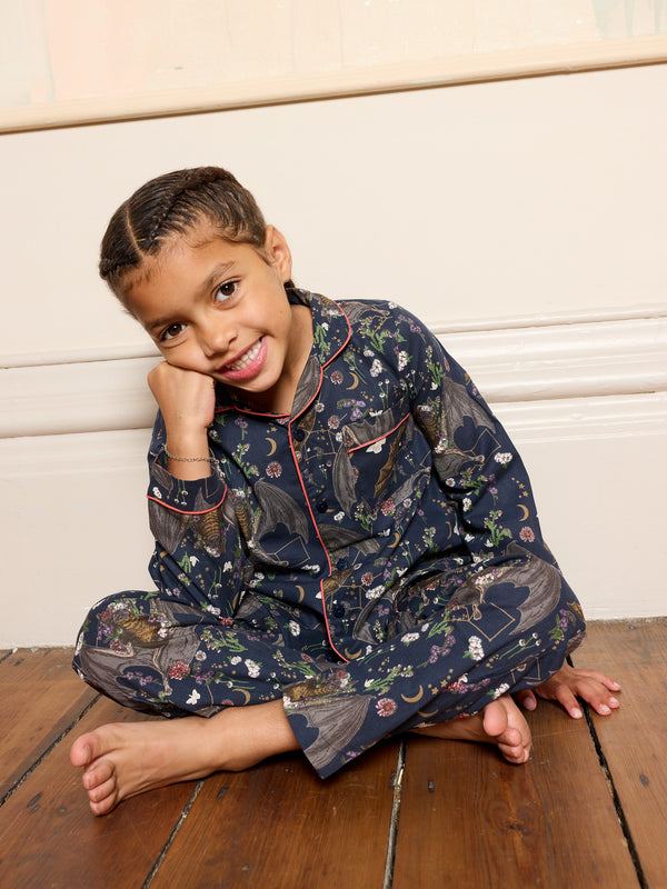 Child wearing a dark grey bat themed pyjama set with a pattern, sitting on a wooden floor.