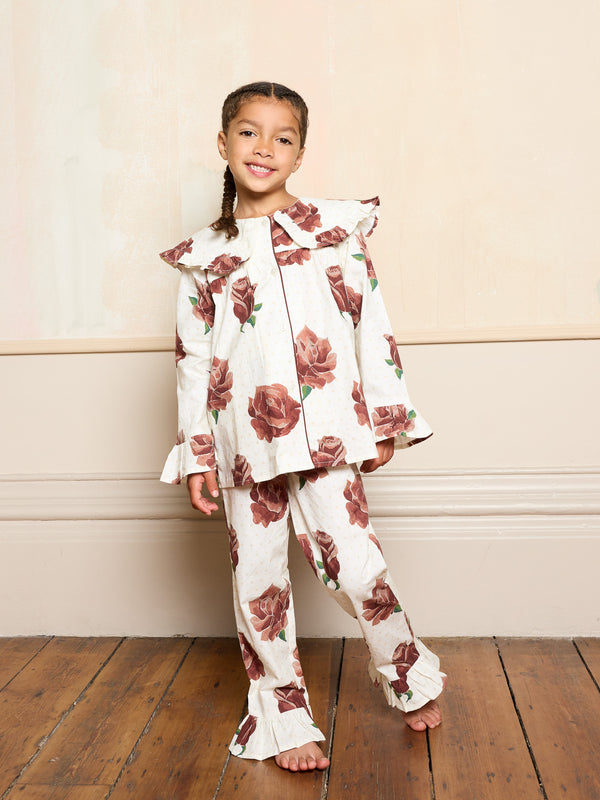 Little girl wearing cream floral pyjama set with a peter pan collar and fluted sleeves standing against a plain wall