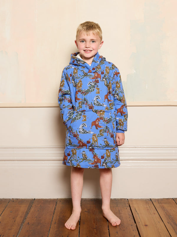 Child wearing a blue animal patterned hooded blanket standing on a wooden floor with a light-coloured wall background
