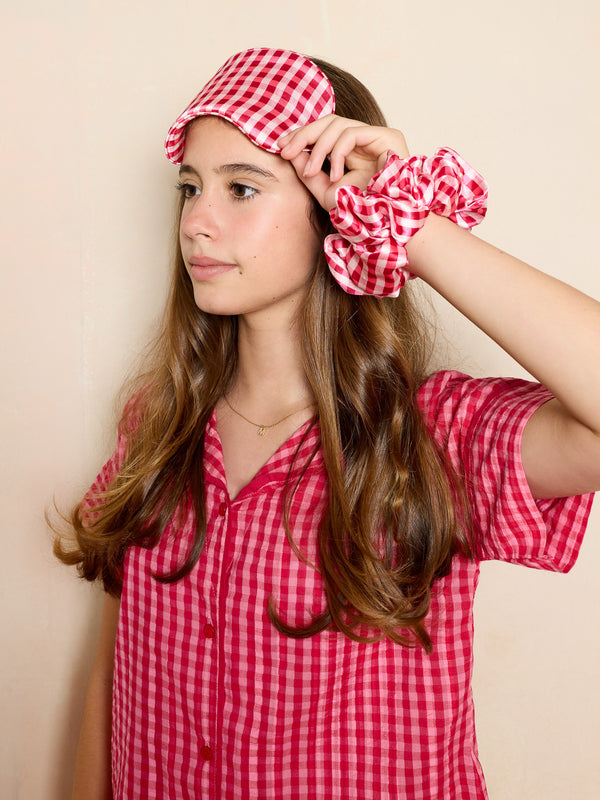 Girl wearing a pink gingham eyemask and scrunchie and pyjama set against a beige background
